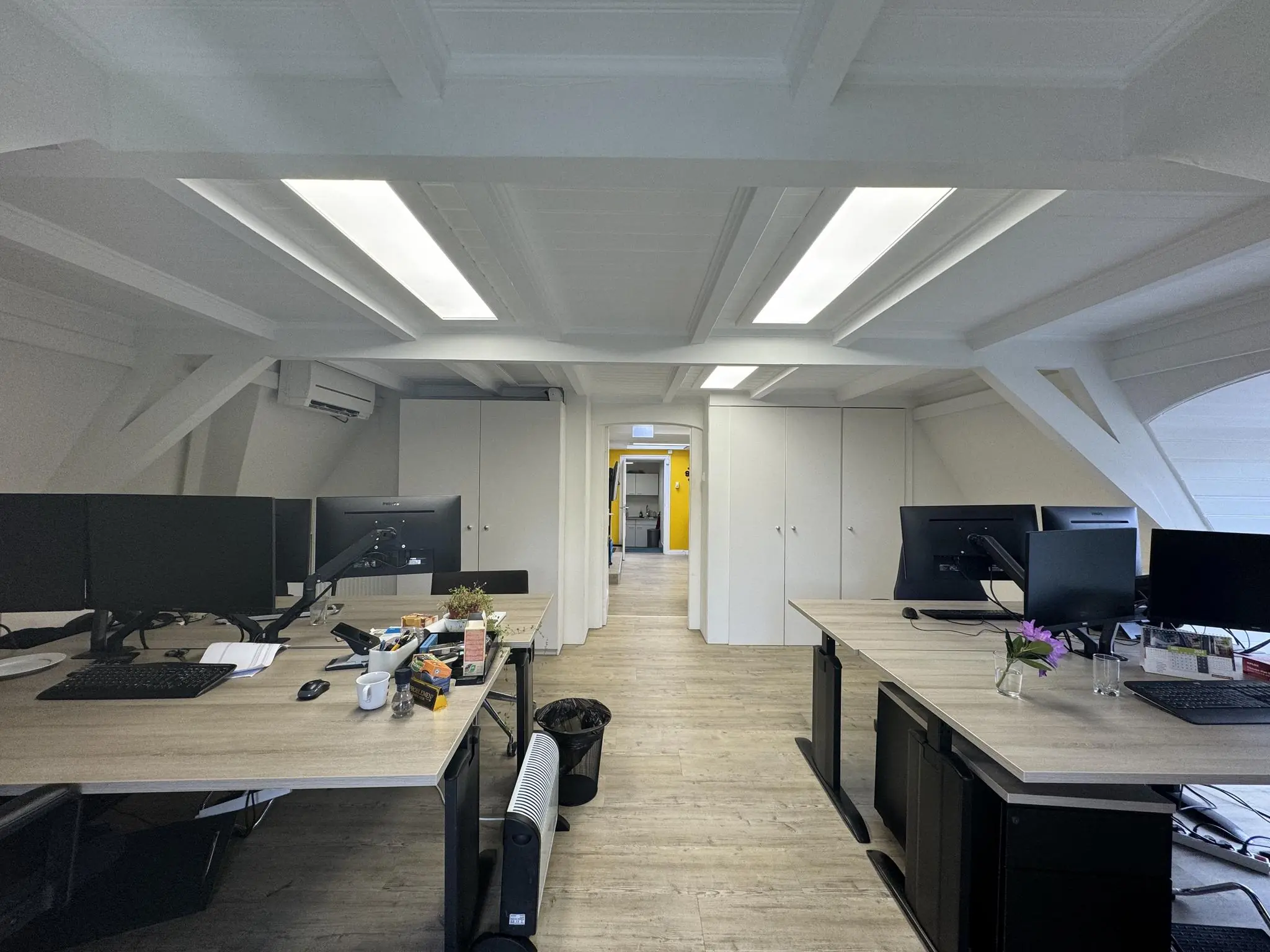 Modern kantoor met houten vloer, meerdere bureaus met computers en zicht op een gang met gele muur achterin.